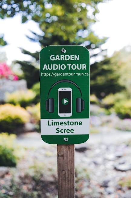 A green signpost with the words 'GARDEN AUDIO TOUR' and a web address, mounted on a wooden stake in a well-maintained garden. The sign features an illustration of headphones and a smartphone with a pl